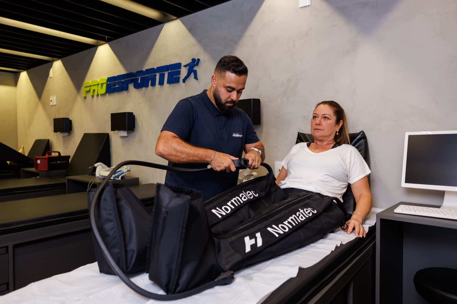 Profissional da Pro Esporte acompanha paciente em sessão de recuperação com botas de compressão, com a marca da clínica na parede ao fundo